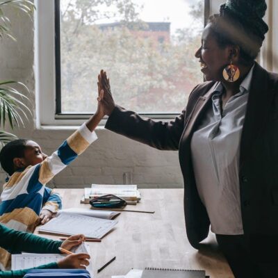 Free Side view of smiling black boy giving high five to teacher while sitting with diverse classmate girl at desk in classroom Stock Photo
